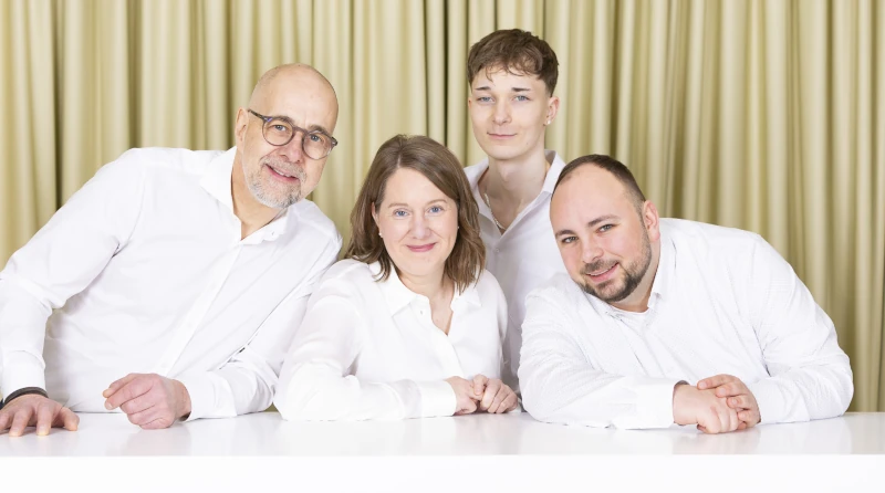 Vier Personen in weißer Kleidung posieren freundlich vor beigem Vorhang am Tisch.